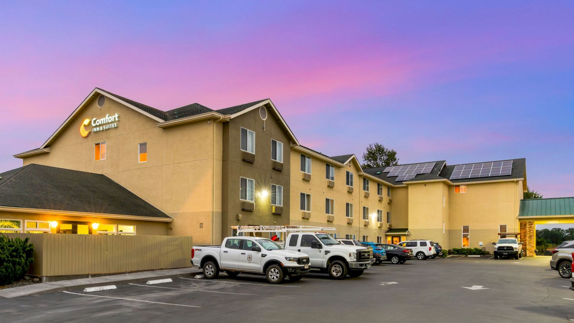 This image shows a Comfort Inn hotel with several white vehicles parked outside, set against a pink and blue evening sky.