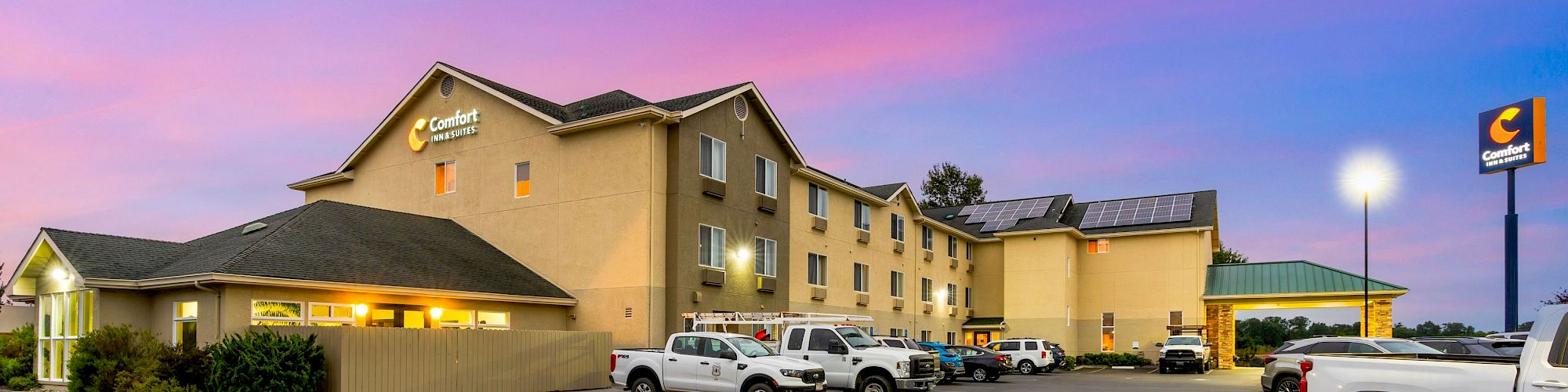 This image shows a Comfort Inn hotel with several white vehicles parked outside, set against a pink and blue evening sky.