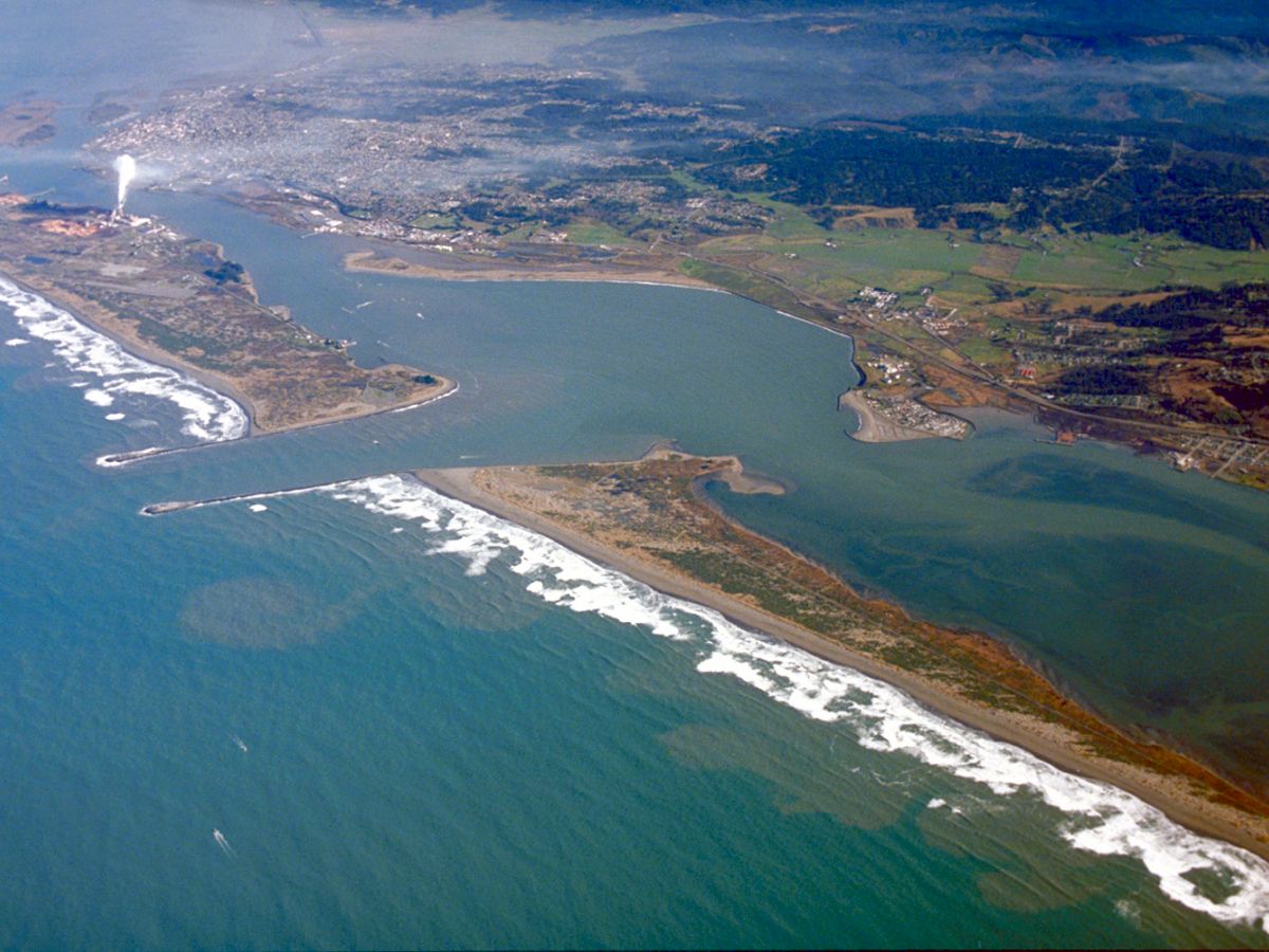 Aerial view of a coastal area featuring a peninsula, surrounding waterways, waves crashing along the shoreline, and a nearby town or city in the background.