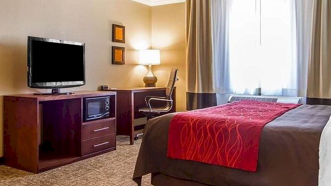 The image shows a hotel room with two beds in separate areas, a TV, a desk with a chair, and bedside lamps with neutral-toned decor.