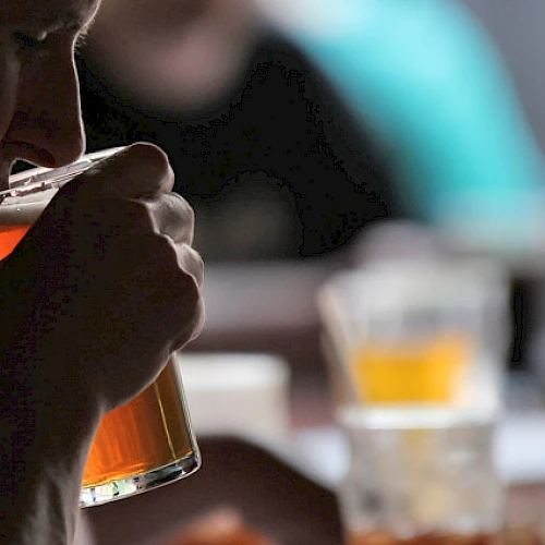A person is drinking beer from a glass in a dimly lit bar or pub, with more drinks visible in the background.