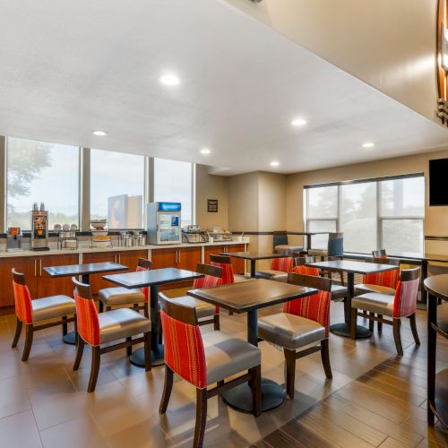 The image shows a modern dining area with several tables and chairs, a counter with beverage dispensers, and a wall-mounted TV. The room is well-lit.