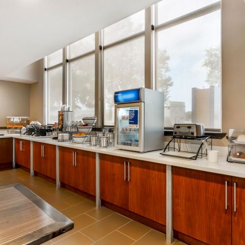 A breakfast buffet setup with coffee machines, cereal dispensers, toasters, and a variety of pastries and bagels, in a well-lit room.