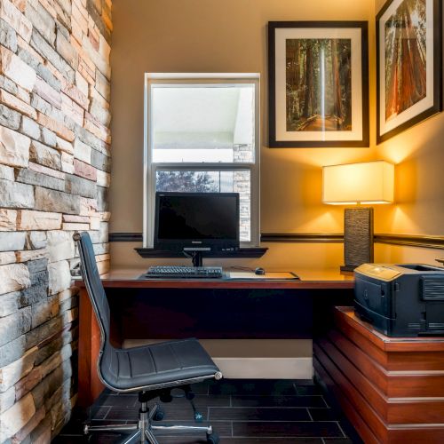 A modern home office setup with a computer on a wooden desk, a black office chair, printer, lamp, and framed pictures on the wall.