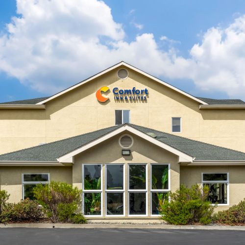 This image shows the exterior of a Comfort Inn & Suites hotel building with a sign on the facade and greenery around the entrance on a sunny day.