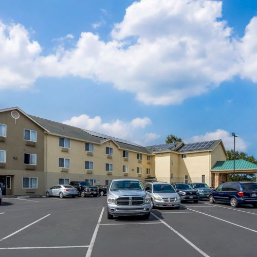 The image shows a beige building with multiple windows, a green-roofed entrance, and a nearly empty parking lot. The sky is blue with clouds.