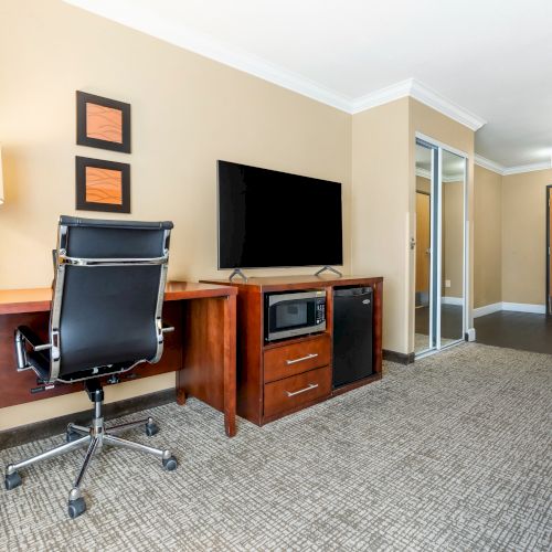 A hotel room with a desk, chair, lamp, TV, and a microwave, with framed art on the wall, door, and a mirrored closet in the background.