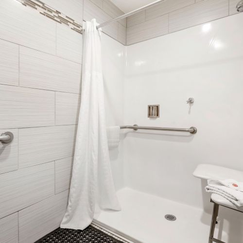 The image displays a modern, accessible shower with a bench, white tiles, grab bars, a shower curtain, and a handheld showerhead.