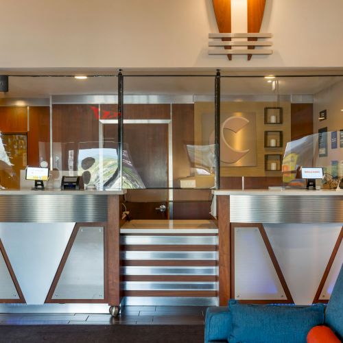 The image shows a modern reception desk with protective screens, information signs, and a waiting area with blue chairs in a well-lit room.