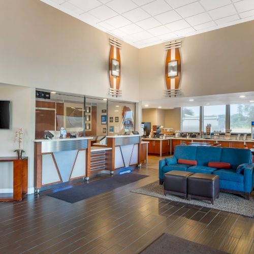 The image shows a modern hotel lobby with a reception desk, seating area, TV, and breakfast bar. The space is well-lit and inviting.