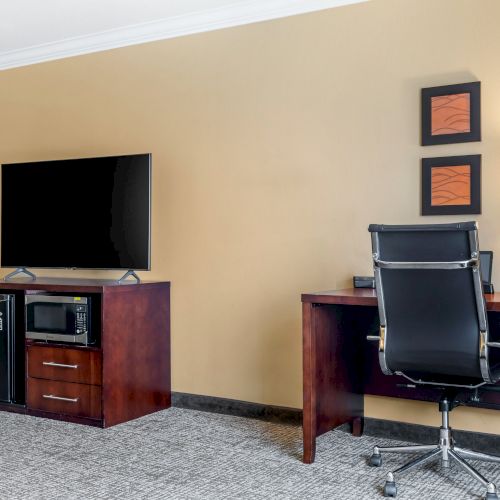 A hotel room features a TV atop a cabinet with a mini-fridge and microwave, a work desk with a lamp, chair, and wall art above the desk ending the sentence.