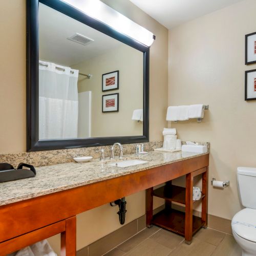 The image shows a well-lit hotel bathroom with a large mirror, sink, toilet, and shower area. The countertop has toiletries and a coffee maker.