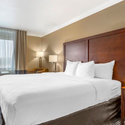 A well-lit hotel room with a large bed, two bedside tables with lamps, a chair by the window, and beige curtains.