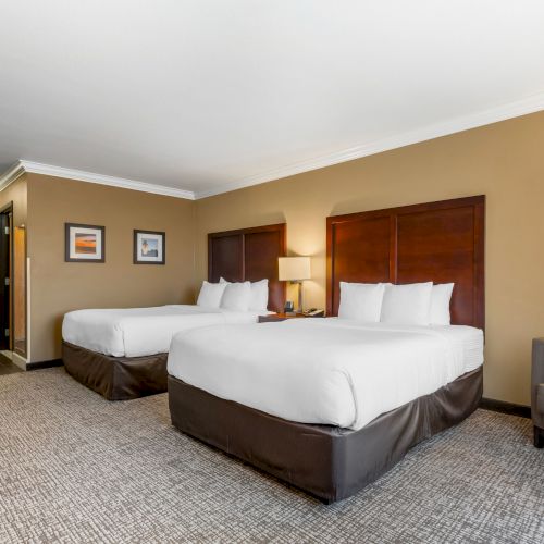 This image shows a well-lit hotel room with two double beds, framed pictures on the wall, and a door leading to the hallway.