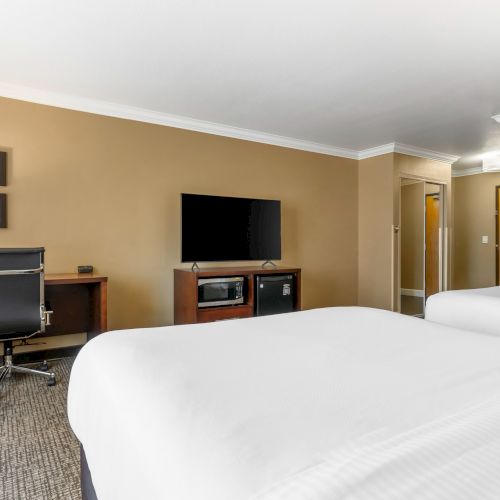 The image shows a hotel room with two beds, a desk with a lamp and chair, a mounted TV, and some framed artwork on the wall.