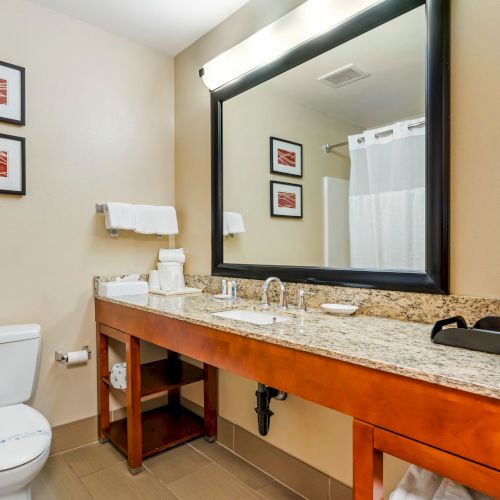 A modern bathroom features a large mirror, a granite countertop sink, a toilet, framed art, and various toiletries neatly arranged on the counter.