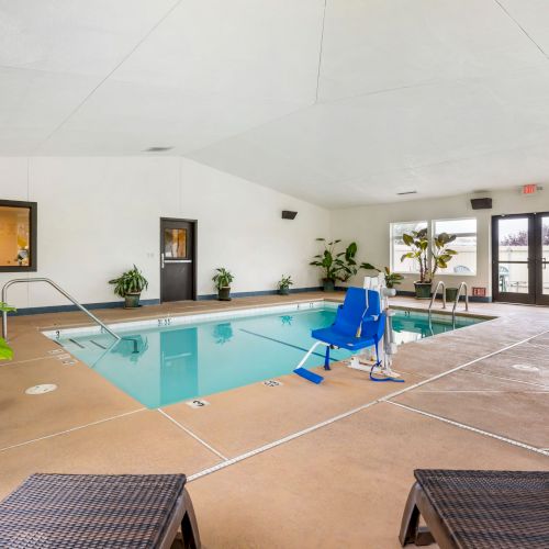 An indoor pool area with lounge chairs, a pool lift, plants, and a seating area by large windows and doors, leading to an outdoor space.