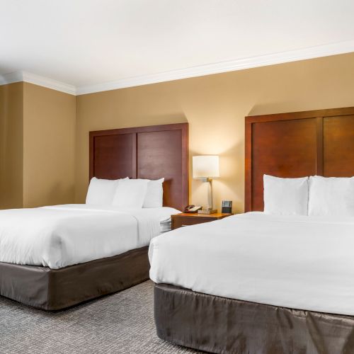 This image shows a hotel room with two double beds, a nightstand with a lamp and phone, and a window with curtains. The walls are beige.