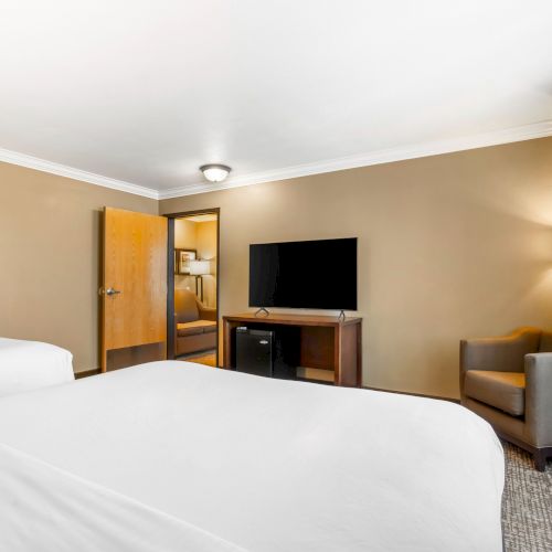 A hotel room with two beds, a TV on a stand, a chair, a lamp, and a partially open door revealing another room.