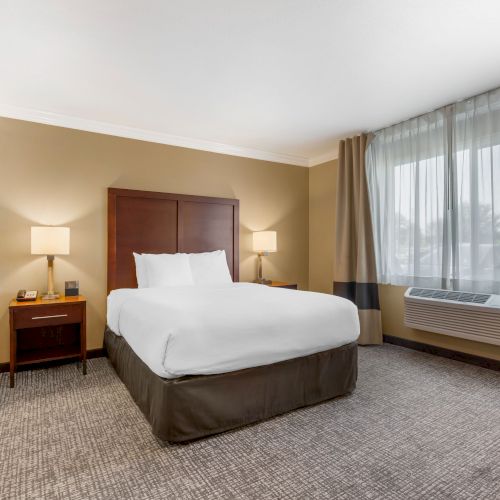 A hotel room with a neatly made bed, two bedside tables with lamps, a window with curtains, and a doorway leading to a bathroom.