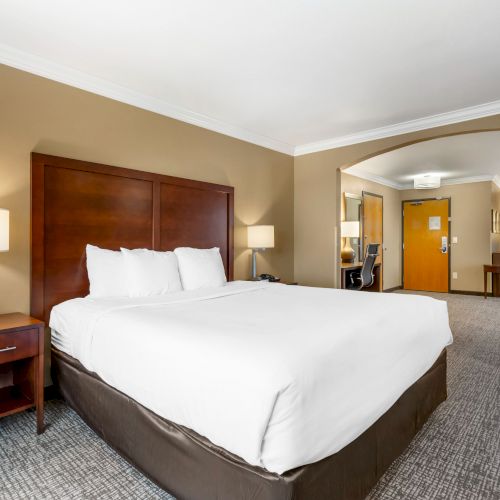 The image shows a hotel room with a large bed, bedside tables with lamps, and a desk in the background. The room is well-lit with a neutral color scheme.