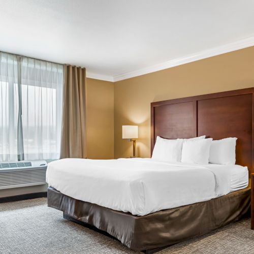 A well-lit hotel room with a large bed, two bedside tables with lamps, an air conditioner under the window, and beige curtains.