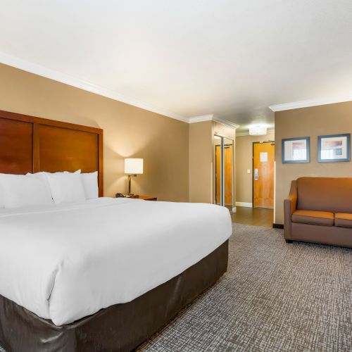 A hotel room with a king-size bed, a brown couch, a bedside lamp, and framed wall art. The room has neutral tones and appears spacious.