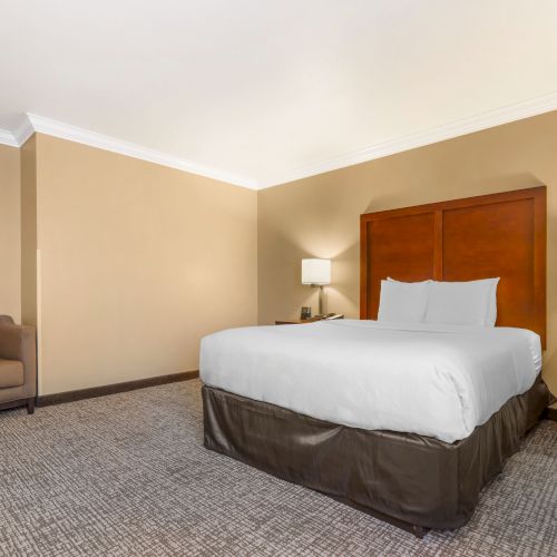 The image shows a bedroom with a double bed, two bedside tables with lamps, neutral walls, and a cushioned chair in the corner.