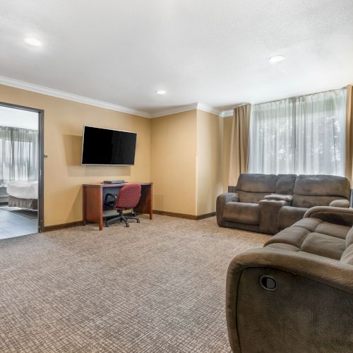 A cozy hotel room with a work desk, TV, couch, and armchairs. The open door leads to a bathroom. The room has beige walls and carpet.