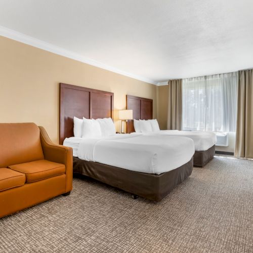 This image shows a hotel room with two double beds, a sofa, a nightstand with a lamp, and a window with curtains illuminating the room.