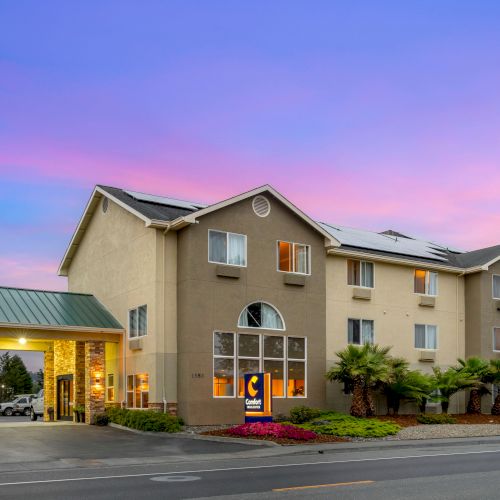 A hotel building with a green roofed entrance area and a sign, captured during sunset with a colorful sky in the background, along a road.