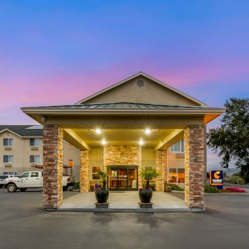 The image depicts the entrance of a hotel with stone pillars, surrounded by a well-lit parking area during sunset, with a tree and hotel sign visible.