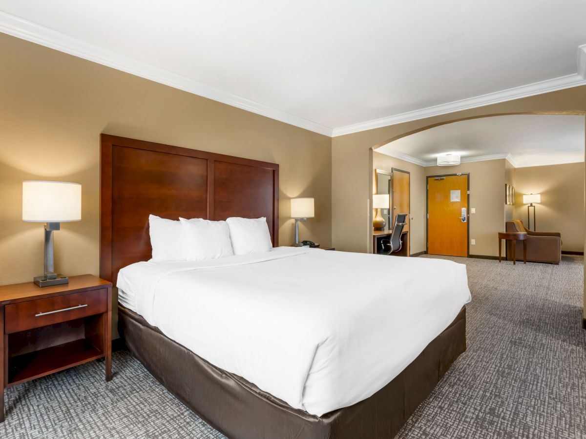 The image shows a hotel room with a large bed, bedside tables with lamps, a desk, and a chair near the door.