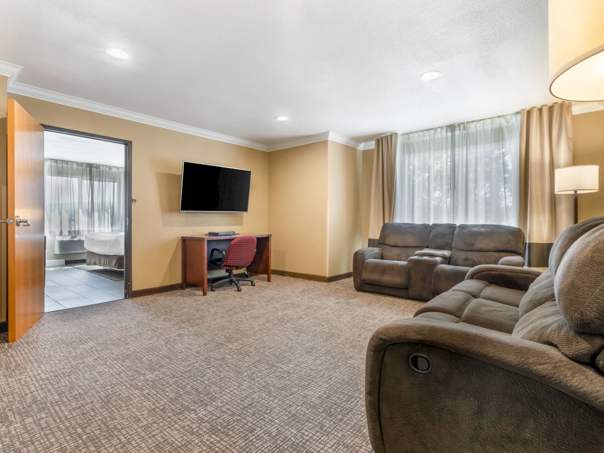 The image shows a cozy living room with a desk, wall-mounted TV, and comfortable seating. A partially open door reveals a bathroom.