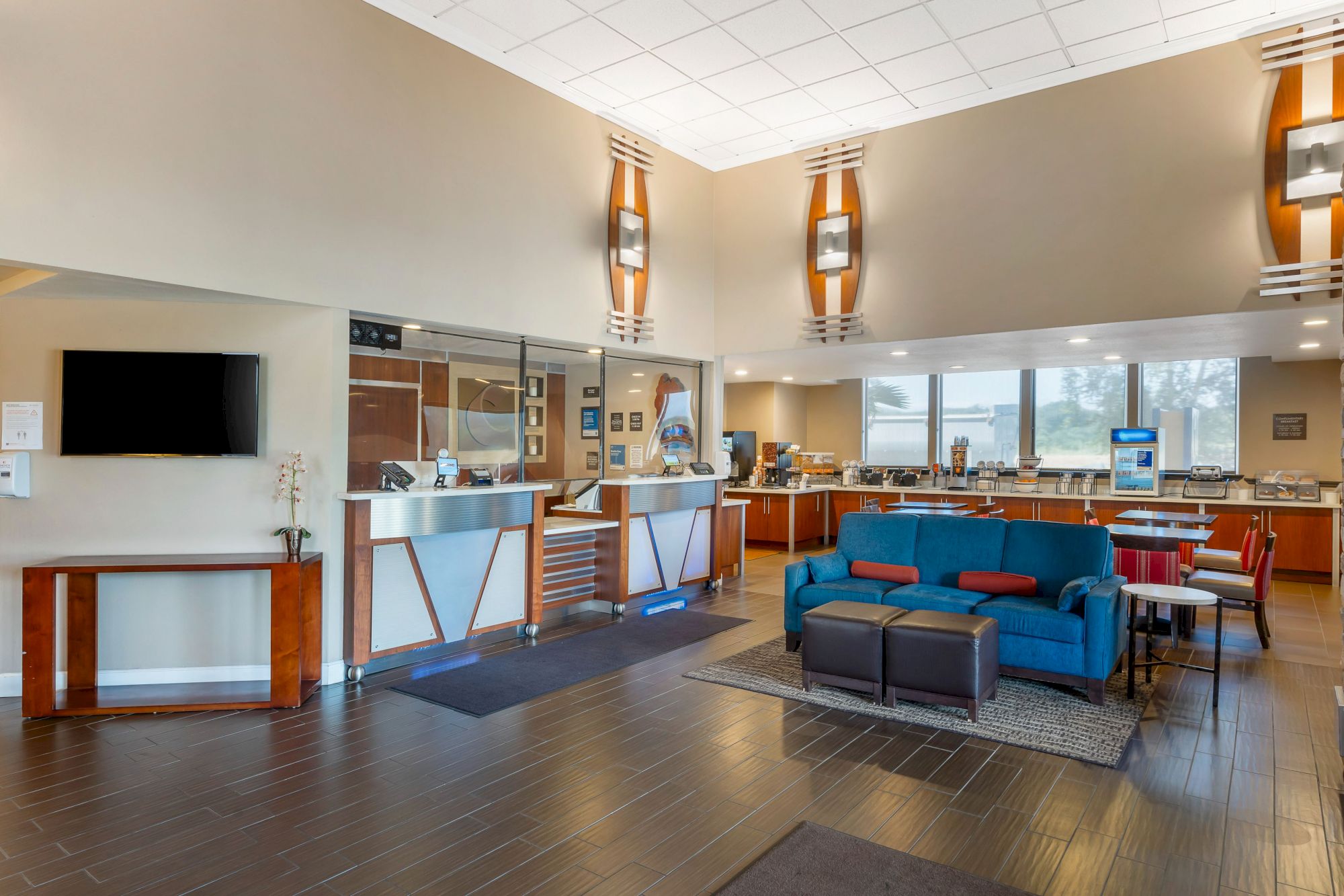 A modern lobby with a seating area, reception desk, TV, and a well-lit breakfast bar in the background, featuring contemporary decor and wooden flooring.