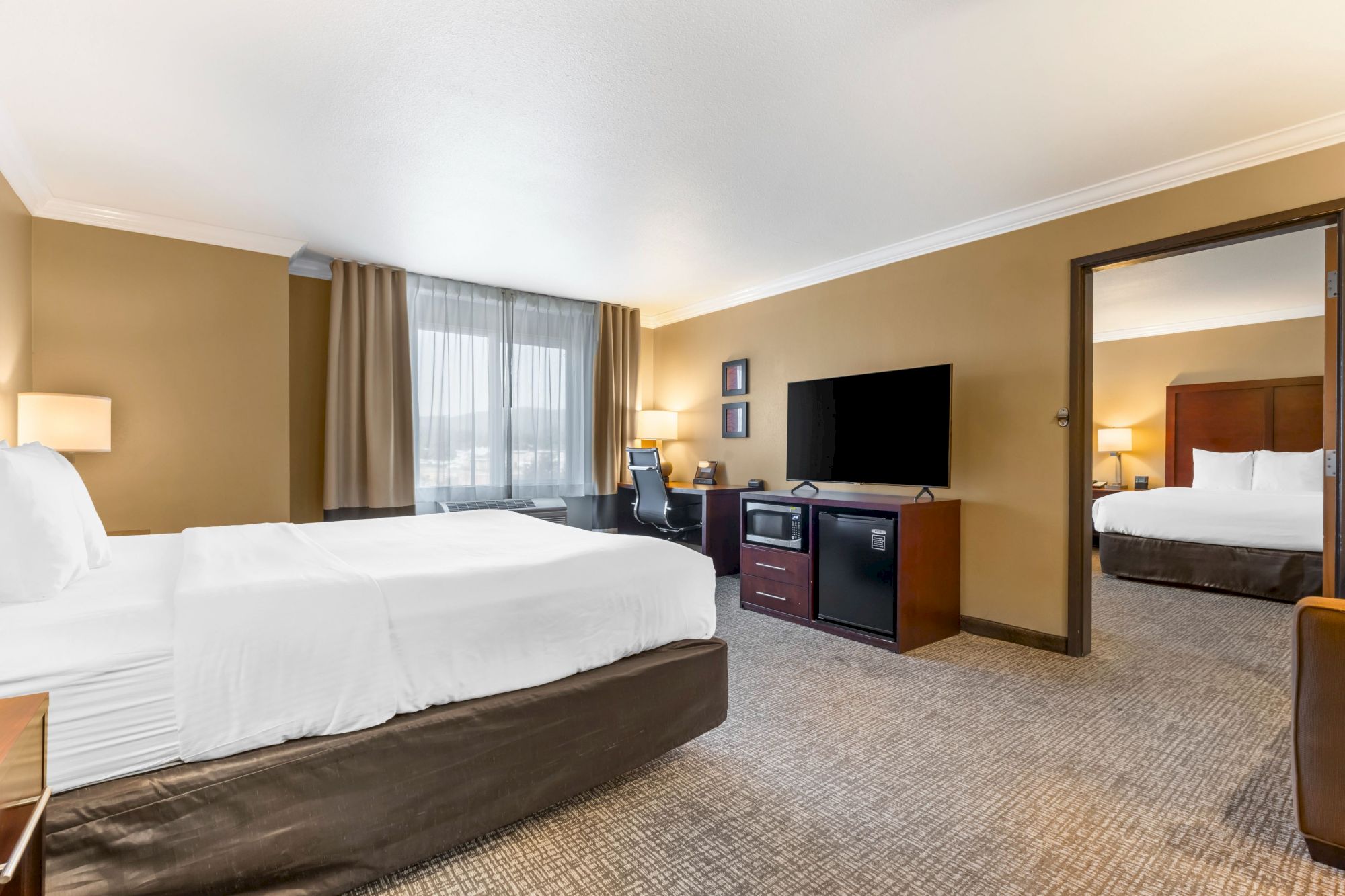 This image shows a hotel suite with two rooms, each containing a bed, a flat-screen TV, a microwave, and modern furnishings in neutral tones.