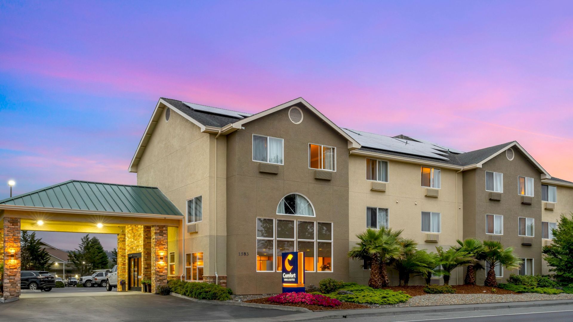 The image shows a three-story hotel building with the Comfort Inn logo, a portico, landscaped garden, and a vibrant sunset sky in the background.