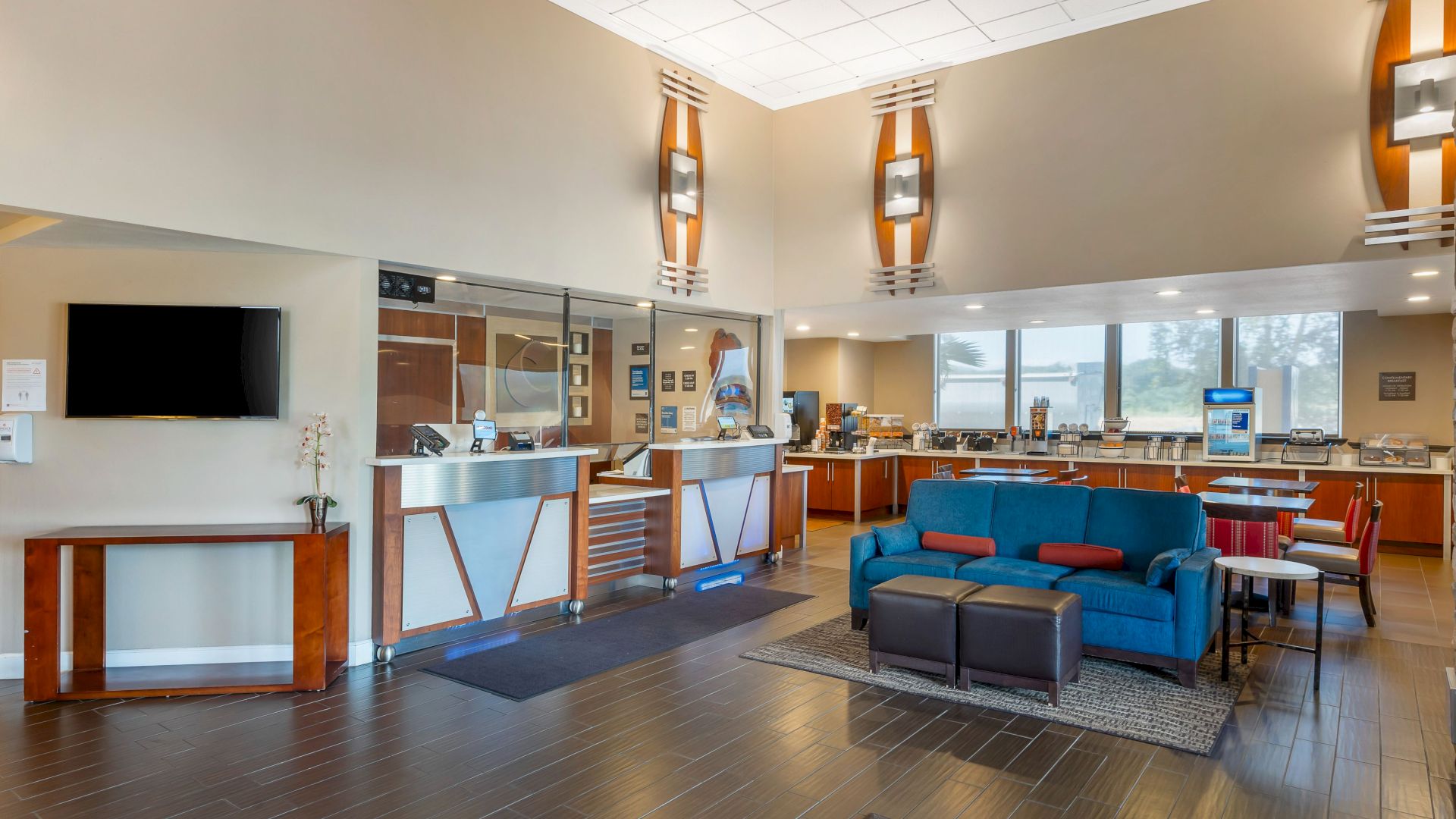 The image shows a modern lobby area with a reception desk, seating, a TV, and a breakfast bar. The space features contemporary decor and ample lighting.