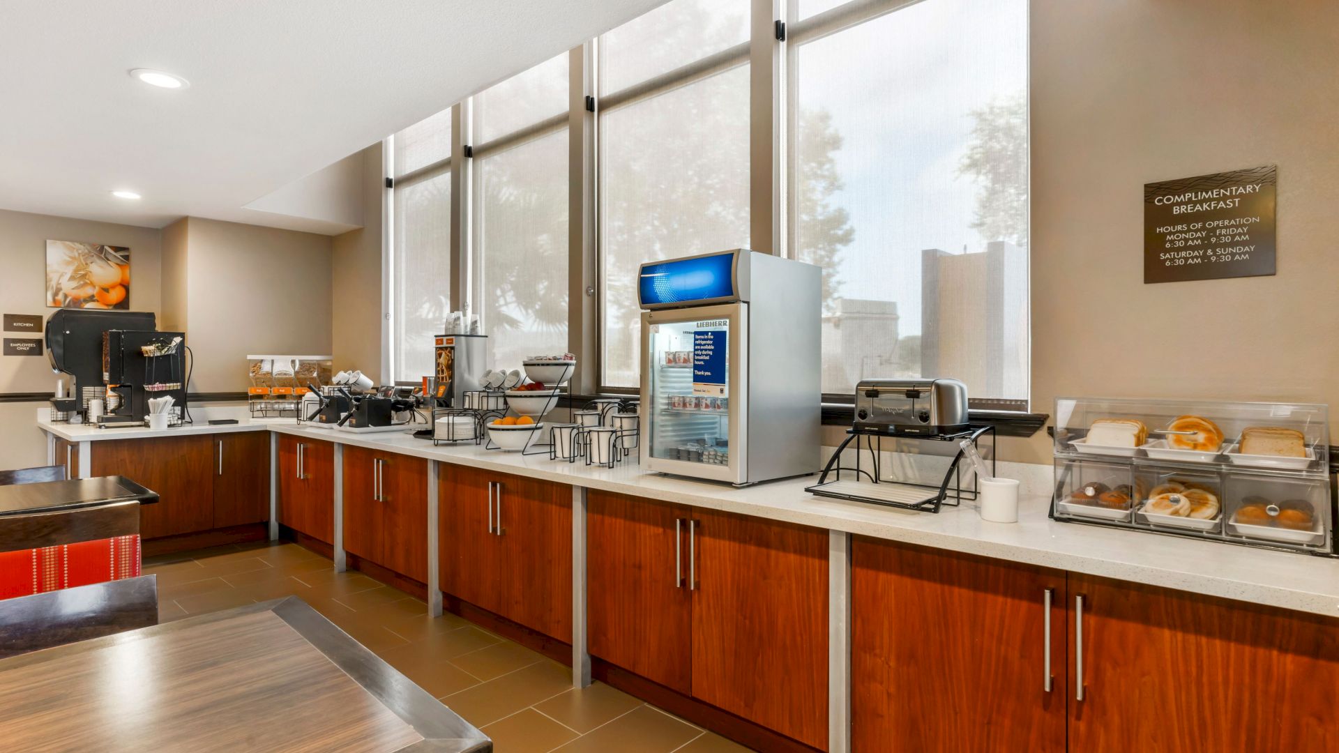 The image shows a hotel breakfast area with a coffee machine, juice dispenser, toaster, and various food items on the countertop.