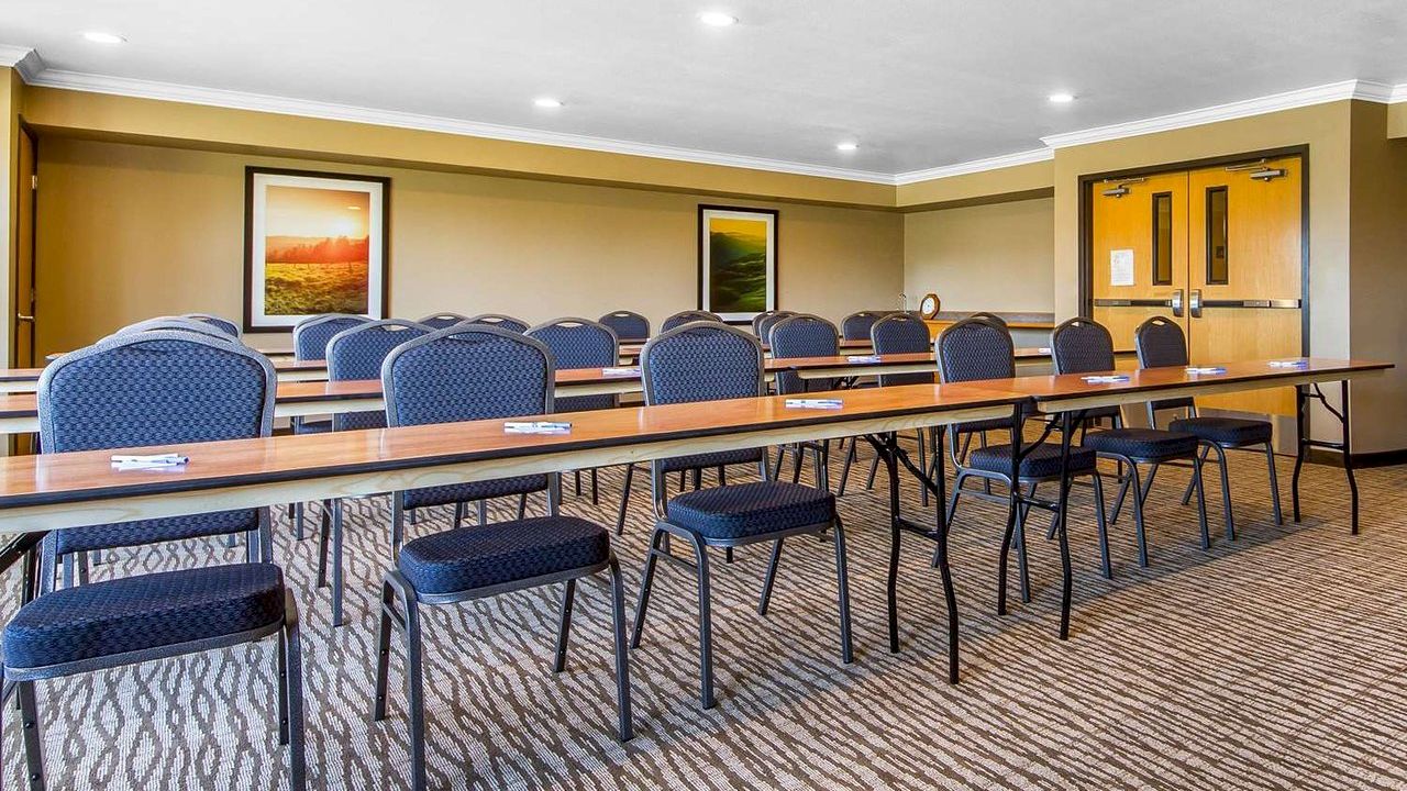 The image shows a conference room with rows of tables and chairs, carpeted floors, framed pictures on the walls, and double doors at the back.