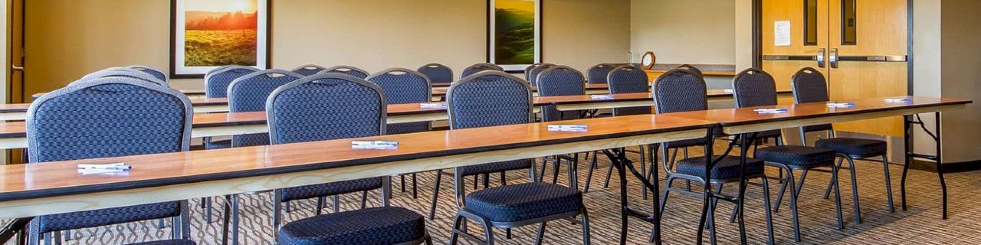 The image shows a conference room with rows of tables and chairs, carpeted floors, framed pictures on the walls, and double doors at the back.