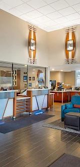 A modern hotel lobby with a reception desk, seating area, and a coffee station, featuring blue couches, warm lighting, and large windows.