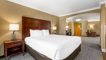 A hotel room with a bed, bedside tables, lamps, a desk, and a door. The room is well-lit and has neutral tones, appearing clean and comfortable.