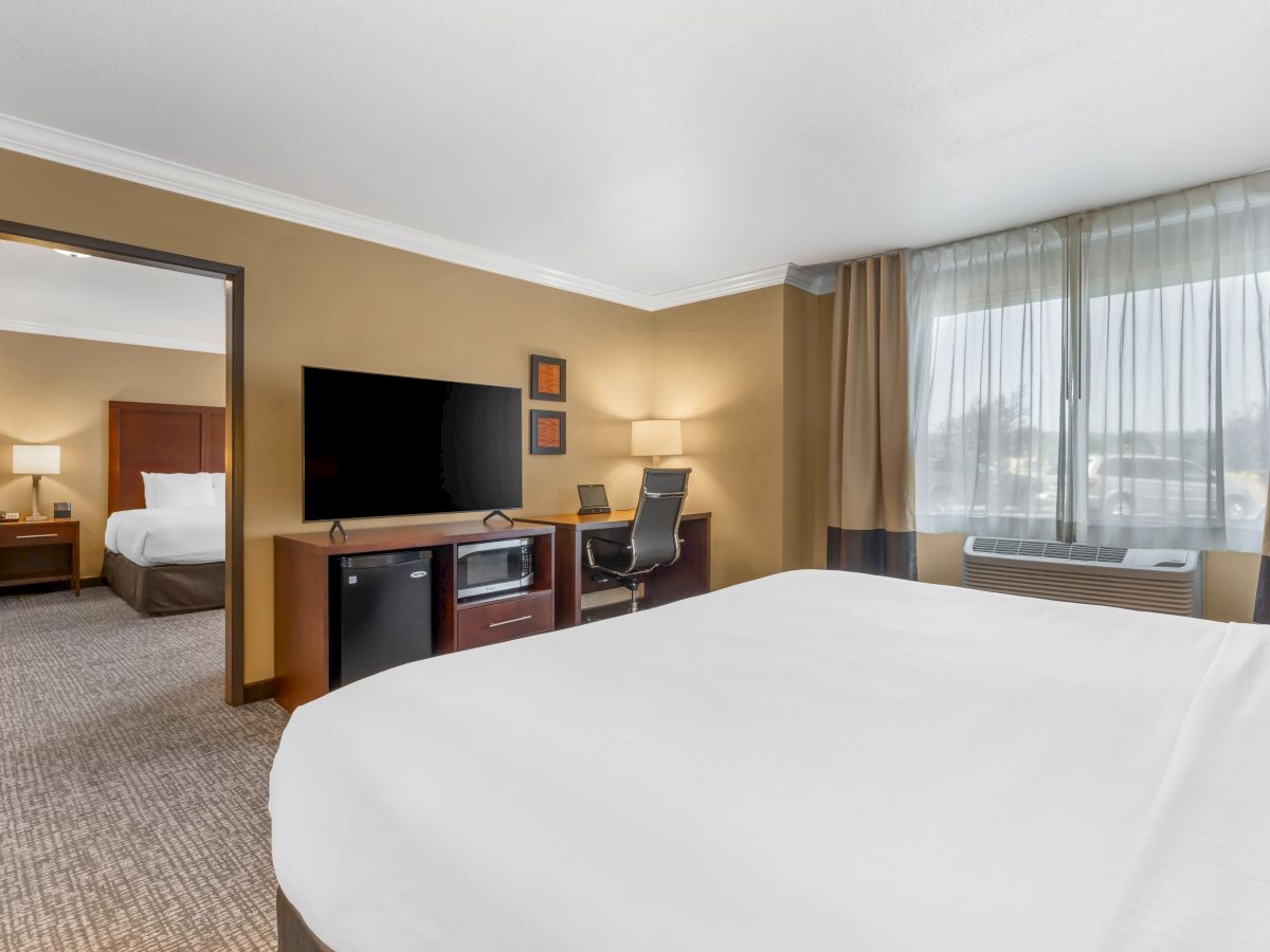 A hotel room with a TV on a desk, a chair, a microwave, and a bed, with another room visible in the background through an open door.