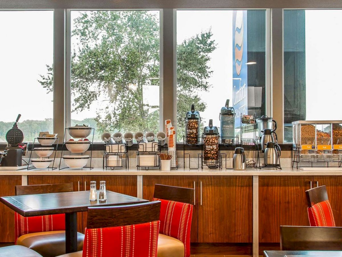 A breakfast buffet setup with various food and drink options, including cereal dispensers, coffee, and a waffle maker, near a window.