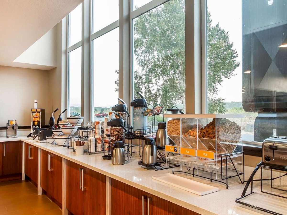 A well-lit breakfast buffet area with various cereals, coffee dispensers, a toaster, and a selection of condiments and snacks is shown.